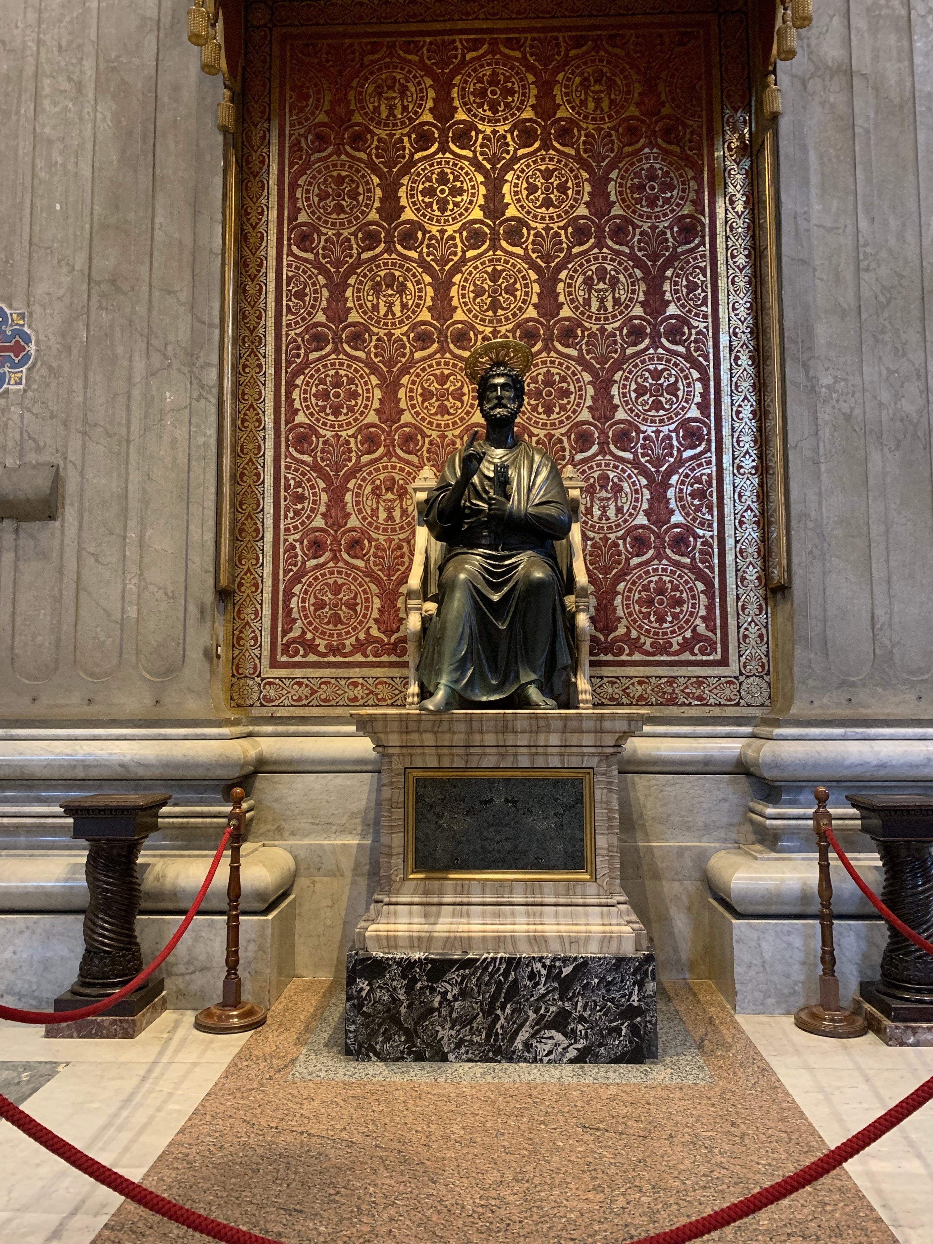Statue of St. Peter in the Vatican it is a dark brown bronze statue that shows a man sitting in a chair with one hand raised.